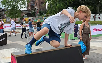 Ein blonder Junge überwindet einen Holzbock mit einer Seitenhocke auf einem Hindernispfad mit Erwachsenen im Hintergrund (Foto)