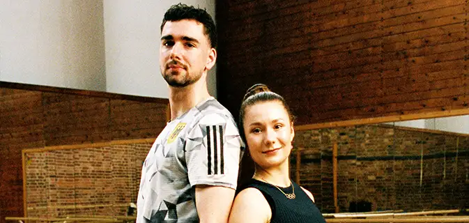 Eiskunstläufer Robert Kunkel und Annika Hocke schauen sich in die Augen beim Tanz auf dem Eis, in dunklen Outfits mit Strasssteinchen und halbtransparenten rötlichen Panelen auf den Oberteilen, mit jeweils einem erhobenen Arm (Foto)